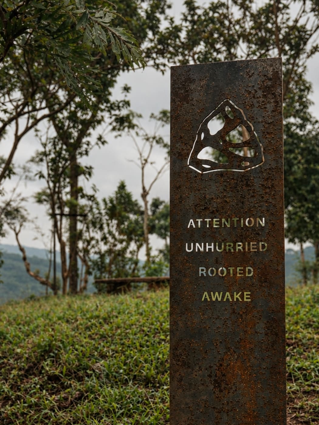 A rusted steel pillar engraved with the Aura mark and the words Attention, Unhurried, Rooted, Awake
