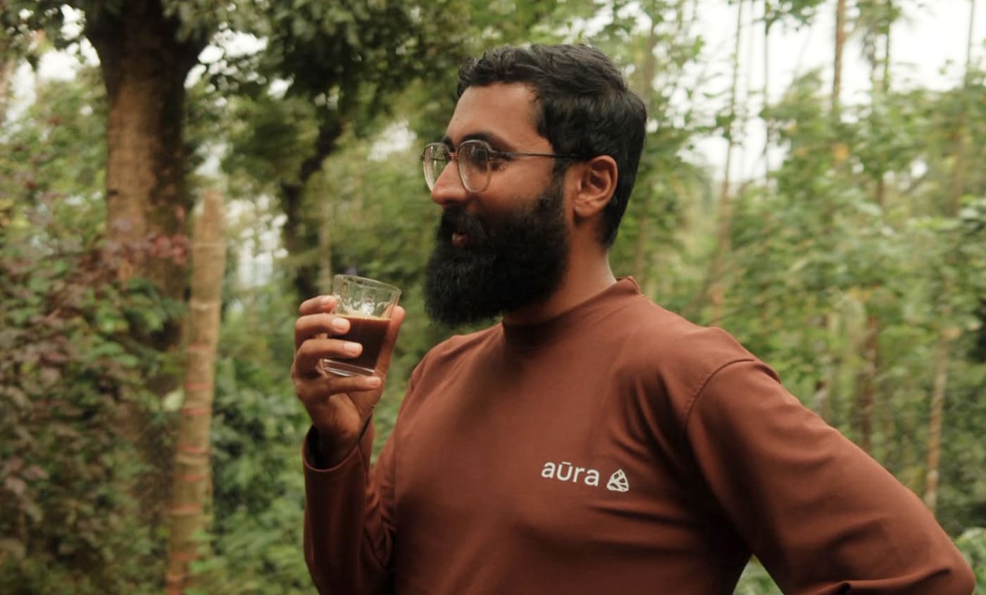 A man in an Aura sweatshirt sipping coffee from a small glass among the trees