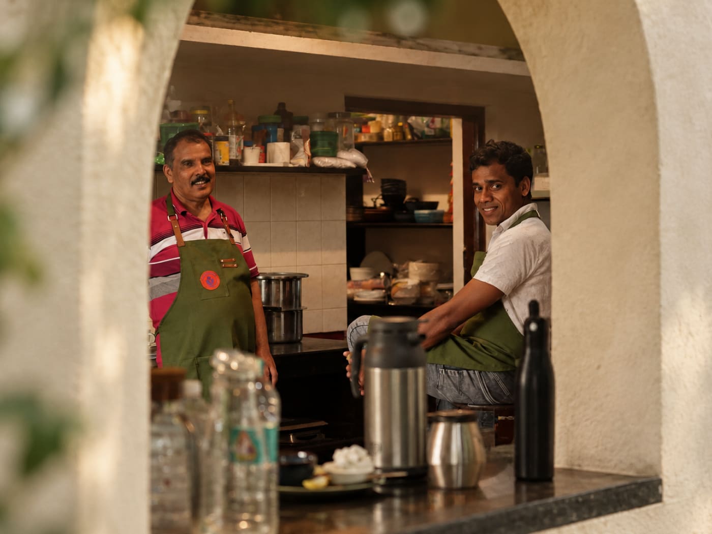 Two cooks in green aprons smiling through an archway into the kitchen