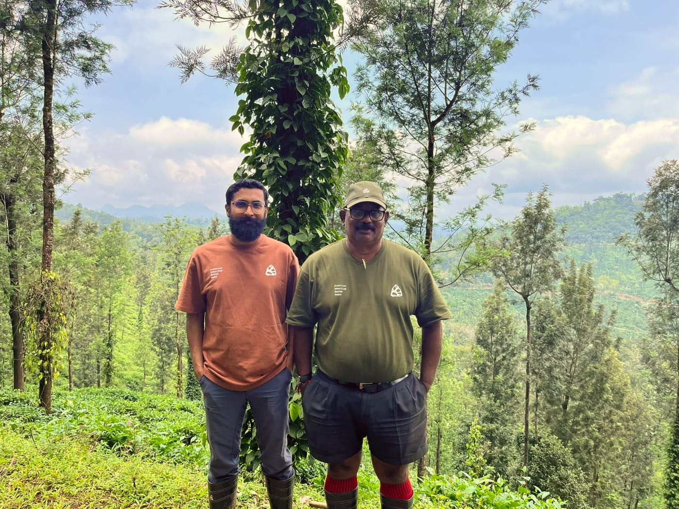 Two men in Aura tops standing among the silver oaks, the tea estate and hills behind them