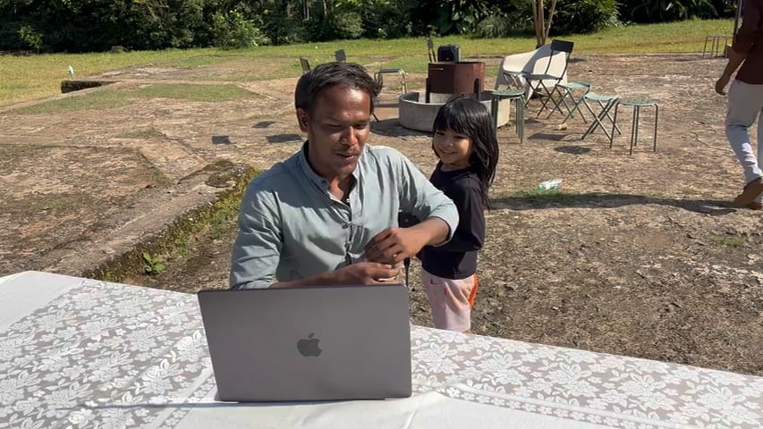 A man at a laptop on an outdoor table, a small girl leaning in beside him