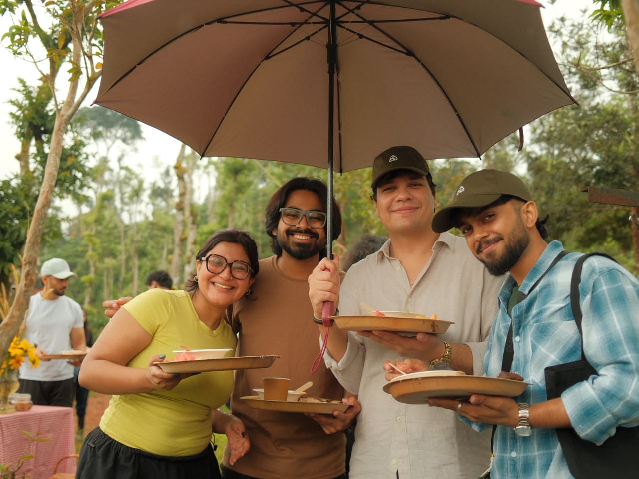 Four friends sharing breakfast plates under a big umbrella in the drizzle