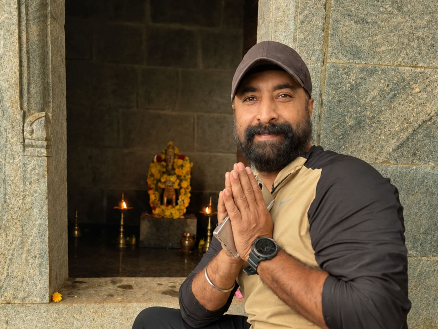 A man in a cap with his hands folded in namaste beside a small stone temple, its deity garlanded and lamps lit