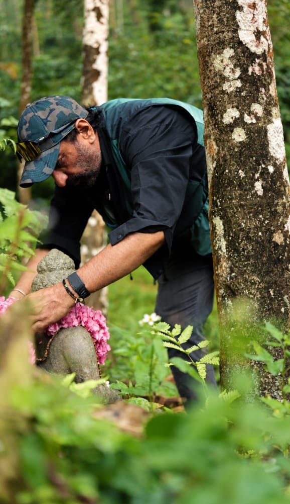 A man garlanding a small stone shrine with pink flowers among the trees